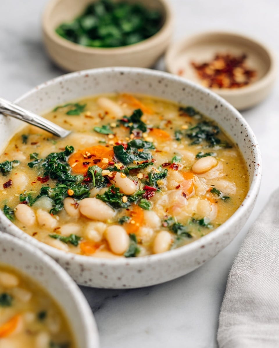 A close-up view of a thick soup in a white speckled bowl, showing chunky white beans and orange carrot slices mixed in a creamy light yellow broth with bits of green leafy herbs on top. The soup has small red chili flakes sprinkled around, adding color contrast. A silver spoon rests inside the bowl on the left side. In the soft-focused background, two small beige bowls hold fresh green herbs and extra chili flakes. The entire setup is placed on a white marbled surface, with a light gray cloth napkin barely visible on the right side. Photo taken with an iphone --ar 4:5 --v 7