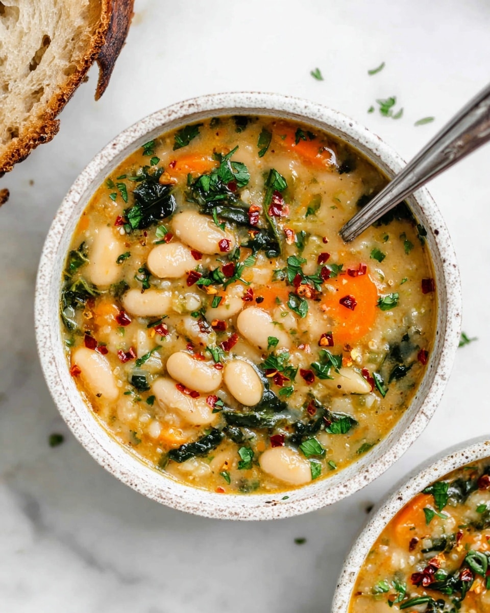 A white speckled bowl filled with thick vegetable soup containing visible layers of large white beans, bright orange carrot slices, and dark green leafy herbs mixed into a creamy, chunky broth layer. The soup is topped with scattered red chili flakes and finely chopped fresh green herbs. A silver spoon rests inside the bowl, slightly dipped in the soup. Part of another similar bowl appears at the bottom of the image, and a torn piece of crusty bread is visible in the top left corner, all placed on a white marbled surface. photo taken with an iphone --ar 4:5 --v 7