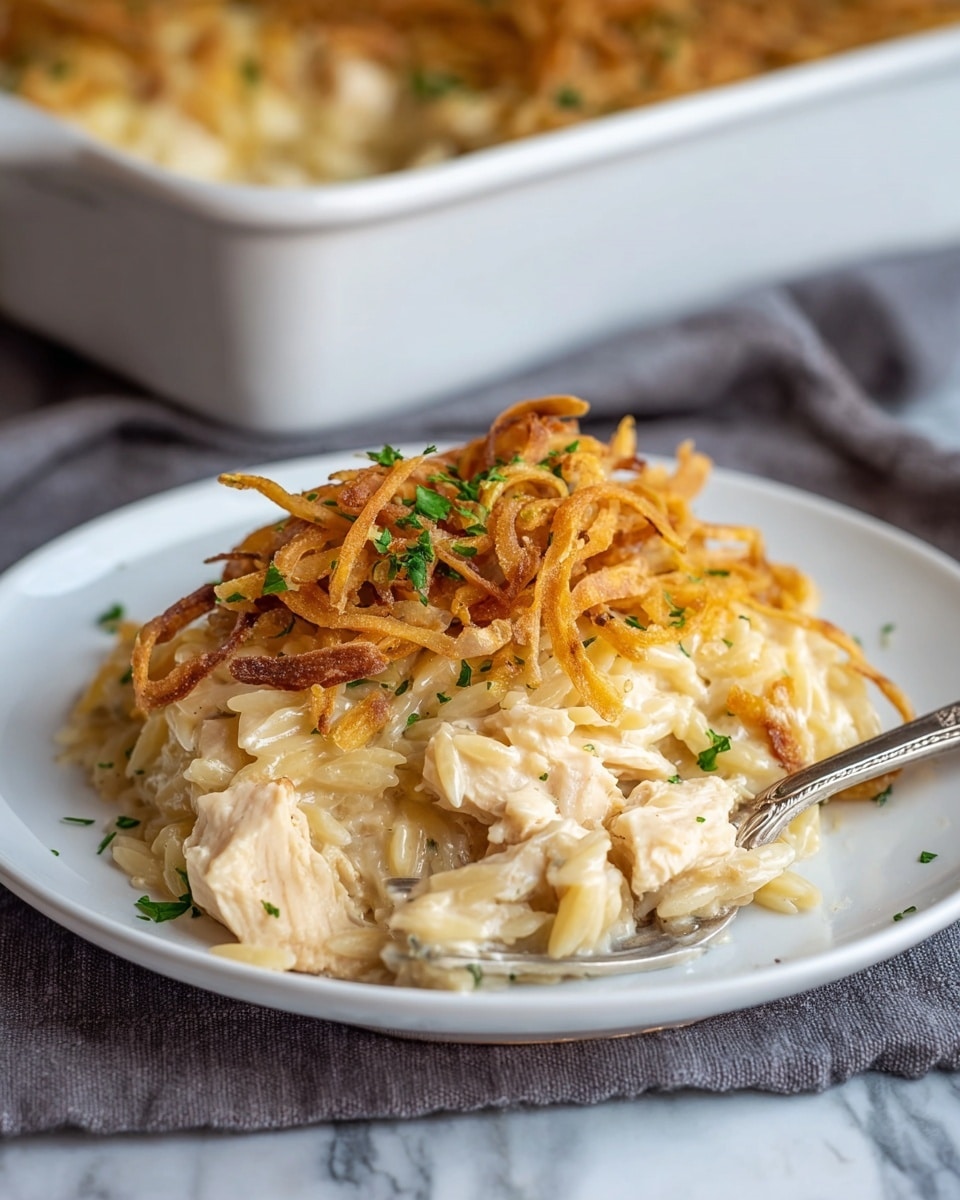 A white plate holds a layered serving of creamy orzo pasta mixed with chunks of white chicken, all coated in a light beige, smooth sauce. On top, a layer of thin, golden-brown crispy fried onions adds texture and color, sprinkled lightly with small pieces of green herbs. The plate is set on a gray cloth over a white marbled surface, and a silver fork rests on the side of the plate. In the blurred background, a white baking dish with the same dish can be seen. photo taken with an iphone --ar 4:5 --v 7