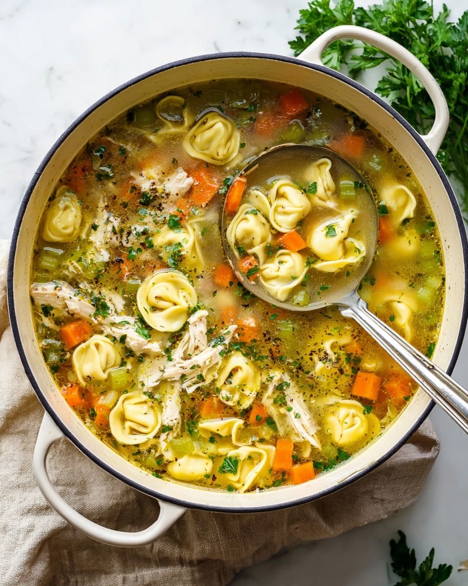 A large white cast iron pot filled with clear broth soup containing three main visible layers: the bottom layer consists of small diced orange carrots and green celery pieces, the middle layer has soft, creamy yellow tortellini pasta and shredded white chicken, and the top layer is sprinkled with fresh green herbs and herbs floating in the broth; a silver ladle is partially submerged in the soup inside the pot, which sits on a light beige cloth on a white marbled surface with some fresh parsley in the background; photo taken with an iphone --ar 4:5 --v 7