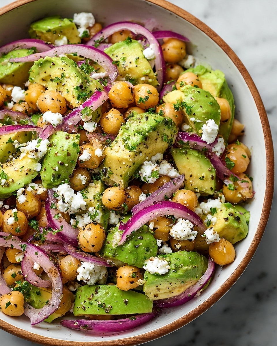 A close-up view of a colorful salad in a white bowl with a wooden rim, showing three main layers: bright green avocado chunks with smooth texture, golden chickpeas that are round and slightly glossy, and thinly sliced purple-red onions with a soft, translucent look, all mixed evenly. Small white dollops of crumbly cheese are scattered on top along with chopped fresh green herbs and coarse black pepper seasoning, giving a fresh and textured appearance. The bowl sits on a white marbled surface. Photo taken with an iphone --ar 4:5 --v 7