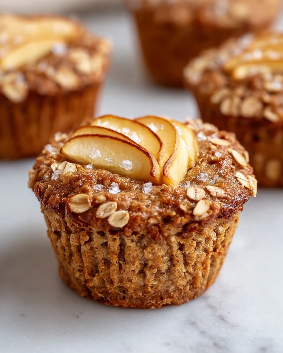The image shows a close-up of a single oat muffin with visible layers and textures. The base layer is a golden brown muffin, surrounded by slightly darker brown ridges from the muffin paper liner. The top layer is covered with rolled oats and cinnamon, giving a rough texture, with thin slices of cooked apple arranged in the center and sprinkled with coarse sugar crystals that glisten under the light. The muffin is placed on a white marbled surface, and two more similar oat muffins are blurred in the background. photo taken with an iphone --ar 4:5 --v 7