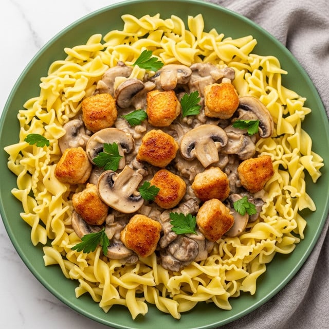 A round green plate holds a base layer of pale yellow egg noodles with wavy edges, spread evenly across the plate. On top of the noodles, there is a creamy sauce with a light beige color, mixed with slices of browned mushrooms and small golden-brown chicken pieces. The chicken pieces have a slightly crispy texture. Fresh green parsley leaves are sprinkled over the sauce, adding bright green color spots. The plate is set on a white marbled surface with a soft cloth nearby. photo taken with an iphone --ar 4:5 --v 7
