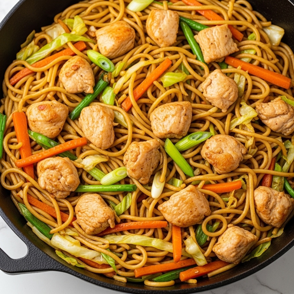 A close-up view of a large black cast iron pan filled with a colorful stir-fry dish featuring long yellowish noodles tangled with thin orange carrot sticks, light green cabbage pieces, and bright green scallion slices. Chunks of cooked chicken, light brown with a slightly glossy texture, are scattered evenly throughout the noodles and vegetables. The ingredients are mixed well, creating a vibrant and textured look with a mix of soft and slightly crunchy elements. The pan sits on a white marbled surface. photo taken with an iphone --ar 4:5 --v 7