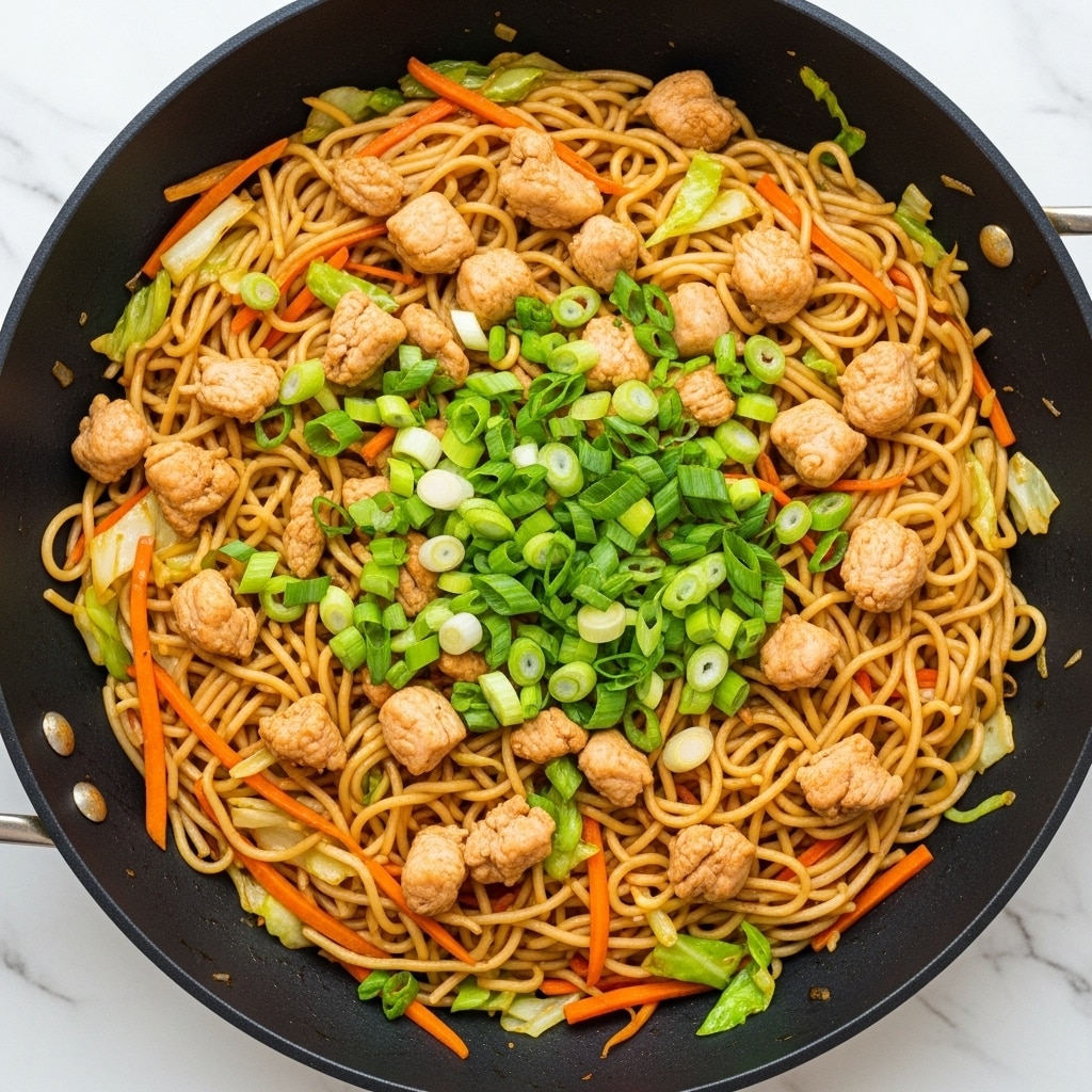 A large black pan filled with stir-fried noodles creates a colorful dish with light golden noodles as the base layer, mixed with thin orange carrot strips and light green cabbage pieces scattered throughout. Small chunks of cooked, light brown chicken are evenly spread in the pan, adding texture and protein. Bright green sliced scallions are sprinkled generously on top, adding fresh color and a slight crunch. The dish sits on a white marbled surface, giving a clean and bright backdrop. photo taken with an iphone --ar 4:5 --v 7