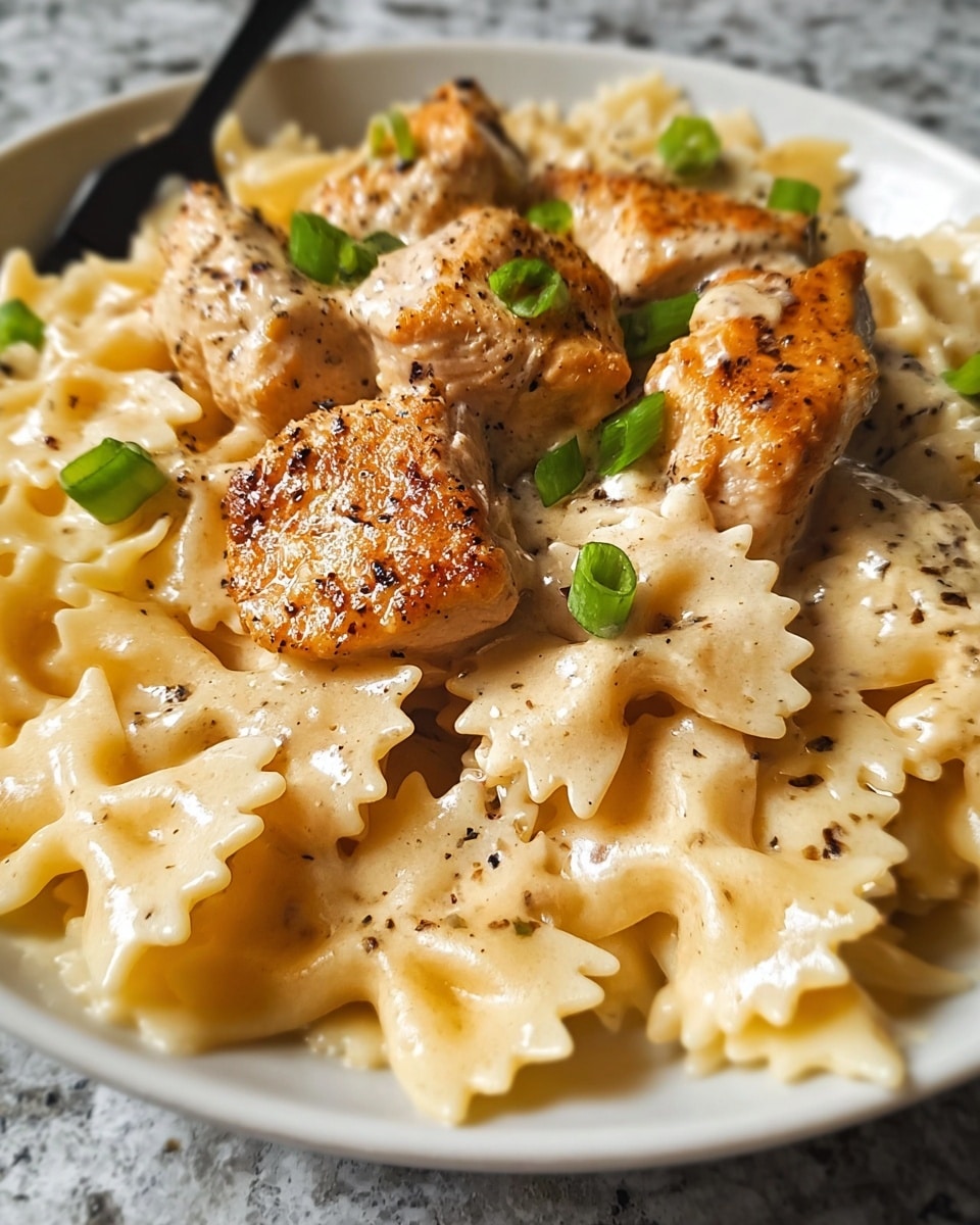 This image shows a close-up of a white plate filled with creamy bowtie pasta covered in a light beige sauce. On top, there are several golden-brown browned chicken pieces with a slightly crispy texture and sprinkled with black pepper. Small green onion slices add a touch of bright green color scattered around the chicken and pasta. The entire dish looks warm, creamy, and freshly made, sitting on a white marbled textured surface. Photo taken with an iphone --ar 4:5 --v 7