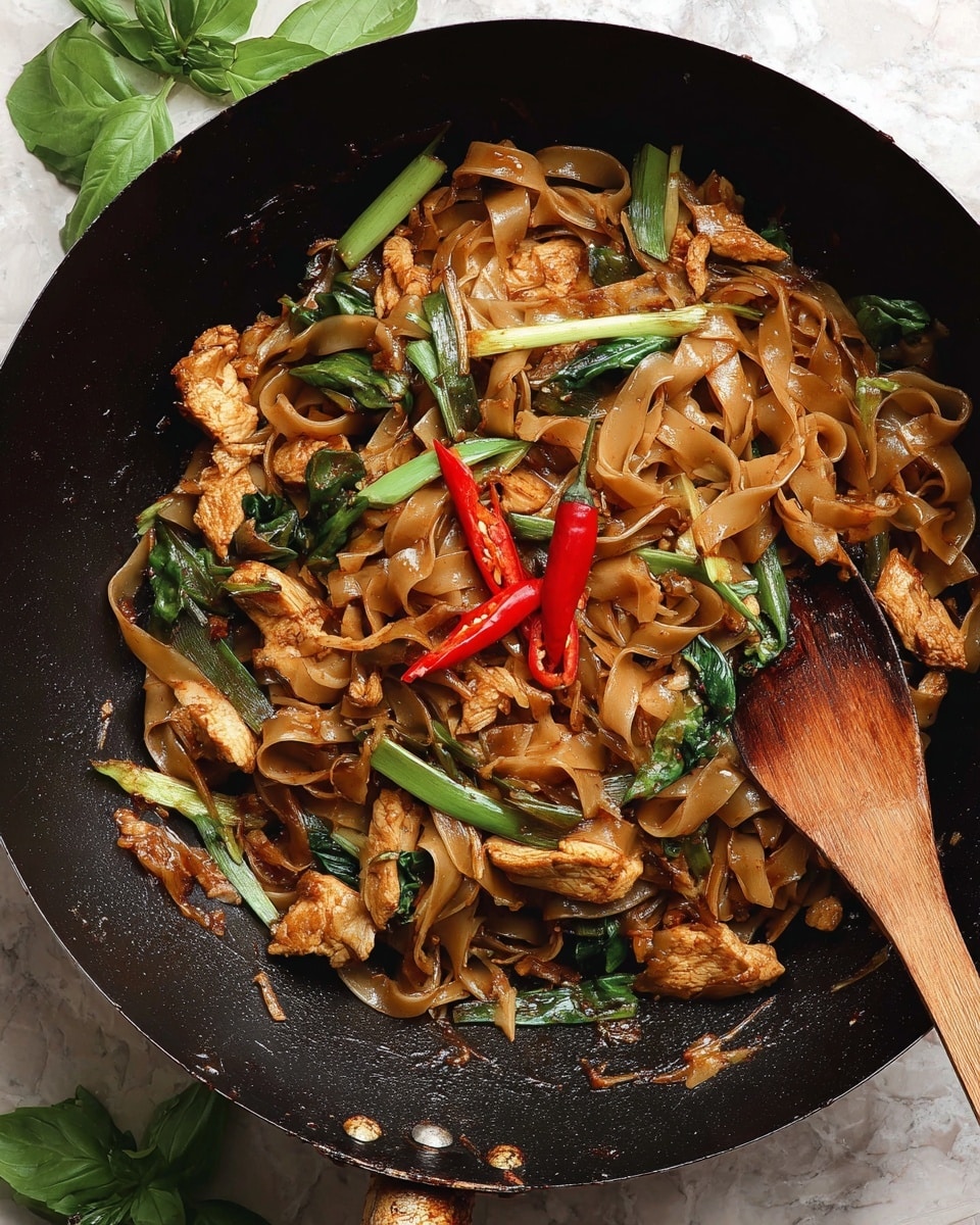 The image shows a black wok filled with a stir-fried noodle dish. The dish has wide, flat brown noodles glistening with sauce as the main layer, mixed with chunks of cooked chicken that are golden brown. There are several pieces of green vegetables including leafy greens and green onion stalks scattered throughout. Two whole red chili peppers are placed on top, with one cut open to show the seeds inside. A wooden spatula rests partly inside the wok, touching the noodles. Fresh green basil leaves are placed on the white marbled surface near the wok, which contrasts with the dark skillet. Photo taken with an iphone --ar 4:5 --v 7