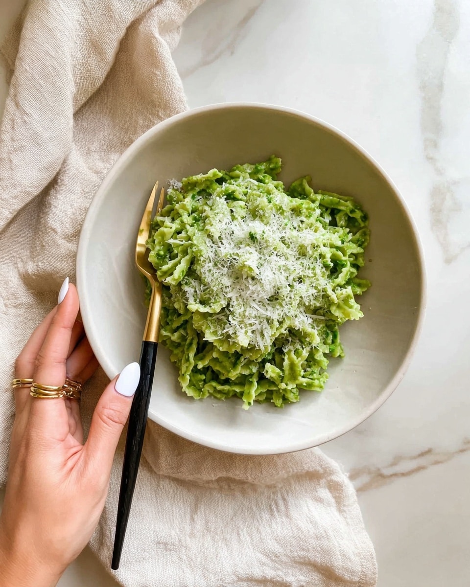 A white bowl holds a serving of green pasta with a ruffled, curly texture, topped with a generous layer of finely grated white cheese. The pasta is piled loosely in the center of the bowl. A gold and black fork rests on the left side inside the bowl. A woman's hand with white painted nails and wearing two gold rings gently holds the edge of the bowl. The bowl is placed on a cream-colored cloth on top of a white marbled surface. Photo taken with an iphone --ar 4:5 --v 7