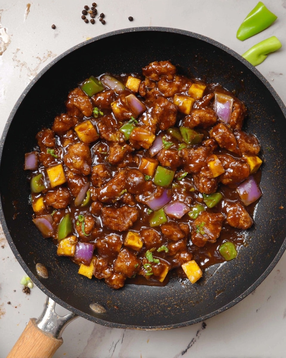 A top view of a black pan filled with small pieces of brown fried chicken covered in a shiny dark brown sauce. Mixed in are yellow cubes of paneer, purple chunks of onion, and green bell pepper pieces, all evenly coated with the thick sauce. The pan has a wooden handle on the left and a metal handle on the right. The background shows a white marbled surface with some scattered pieces of green bell pepper and black peppercorns. photo taken with an iphone --ar 4:5 --v 7