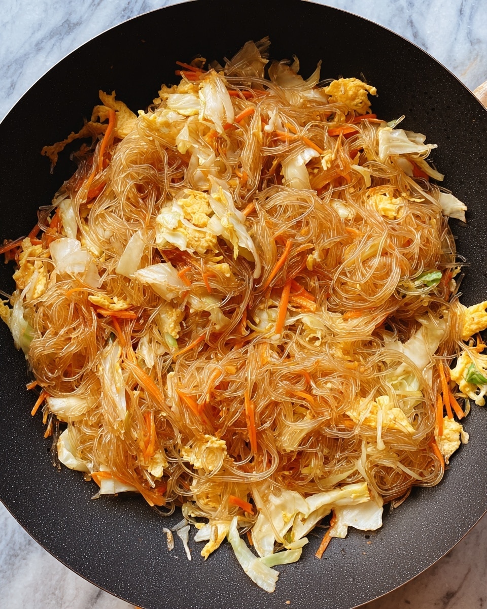 The image shows a wok filled with stir-fried glass noodles mixed with light brown and translucent thin noodles, shredded white cabbage, soft scrambled yellow eggs, and thin orange carrot strips. The ingredients are evenly mixed, showing a combination of smooth, slightly shiny noodle strands and soft, cooked vegetable pieces with some browned edges. The wok is black, contrasting with the light colors of the food inside. The background is a white marbled texture. photo taken with an iphone --ar 4:5 --v 7