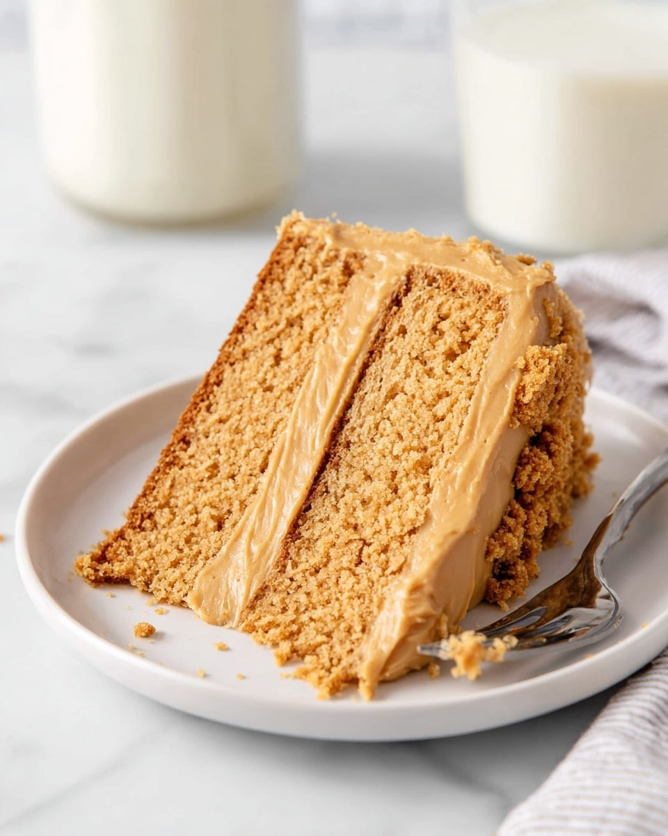 A two-layer slice of light brown cake sits on a white plate, each fluffy cake layer separated by a medium-thick layer of smooth, creamy caramel-colored frosting. The outside of the slice is evenly coated with the same frosting, with small crumbled bits of cake pressed into the back edge. The plate is on a white marbled surface, with a silver fork placed nearby and a glass of white milk blurred softly in the background. Photo taken with an iphone --ar 4:5 --v 7