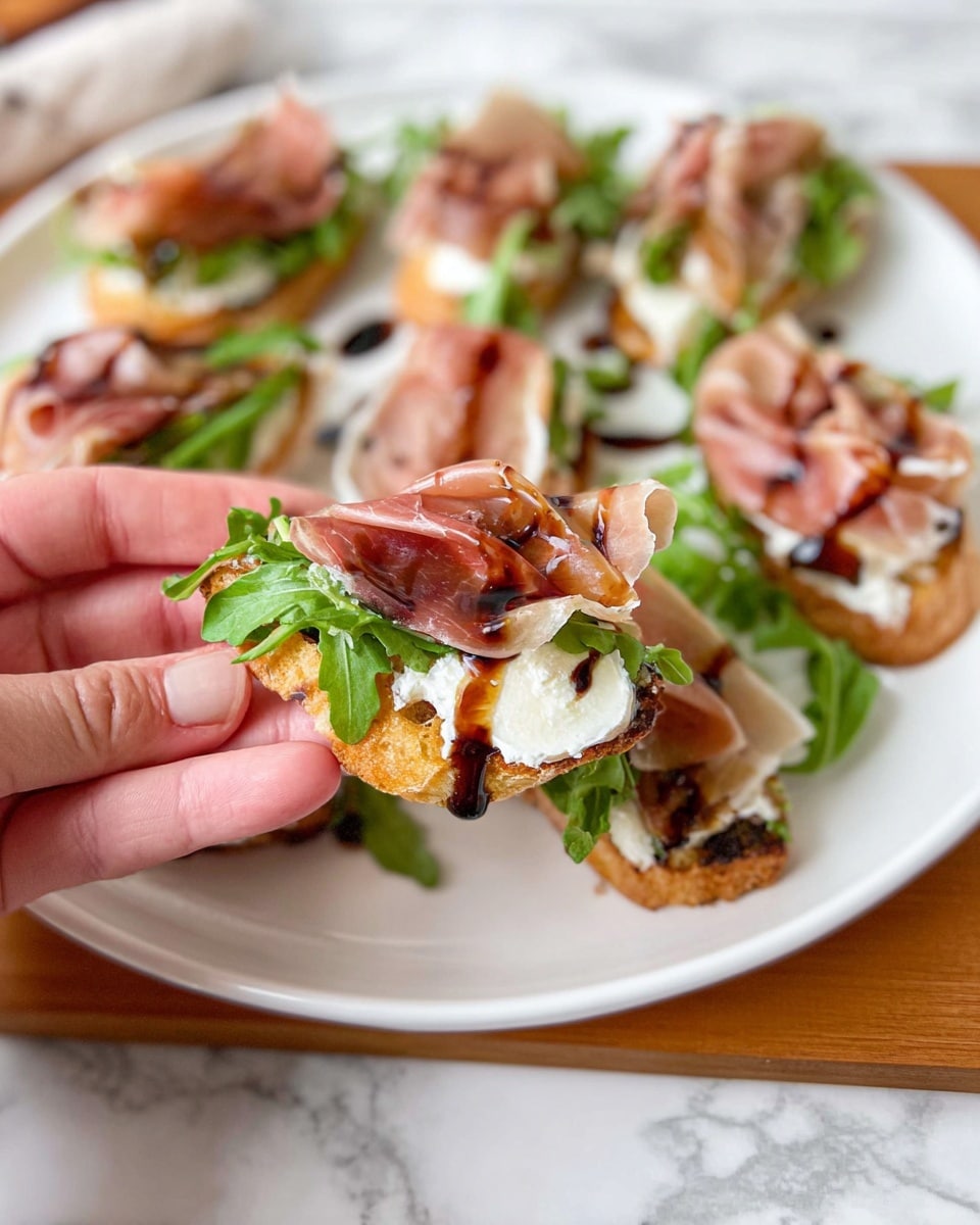 The image shows a white plate with several small toasted bread pieces topped with layers: a creamy white spread at the base, fresh bright green arugula leaves, thin slices of light pink prosciutto, and a drizzle of dark brown balsamic glaze over the top. In the foreground, a woman's hand is holding one piece of the toast, showing the layers clearly with the bread's crisp texture, the soft spread, leafy greens, delicate prosciutto folds, and shiny glaze. The plate is placed on a wooden board over a white marbled surface. photo taken with an iphone --ar 4:5 --v 7