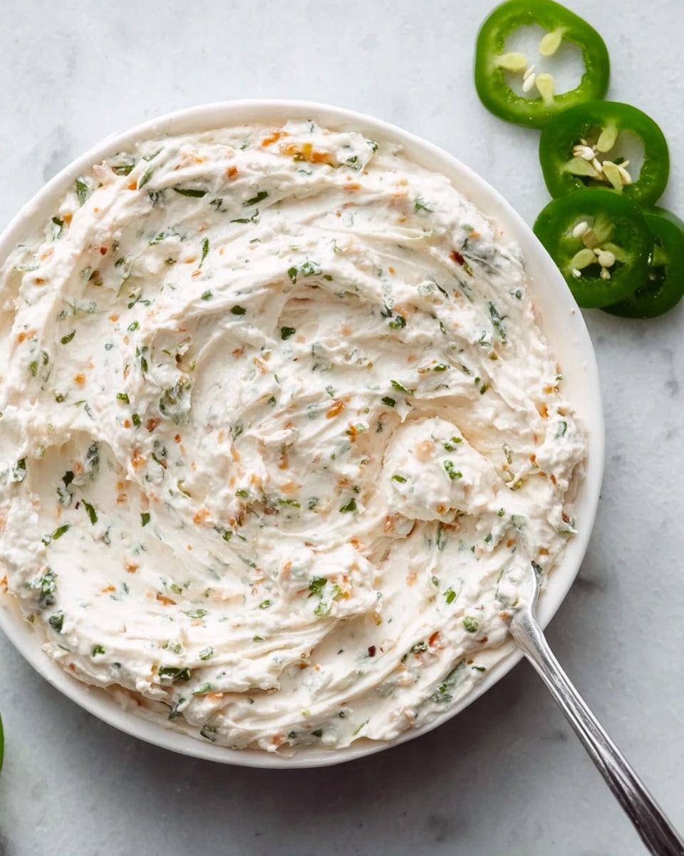 A close-up view of a white round plate filled with a creamy, thick dip that has a slightly whipped texture. The dip is pale off-white with small pieces of green herbs and tiny orange bits mixed throughout, creating a speckled effect. The dip surface shows gentle swirls and folds, with a silver spoon partially dipped into it on the right side. Around the plate, there are some sliced green jalapeño peppers on a white marbled surface. Photo taken with an iphone --ar 4:5 --v 7