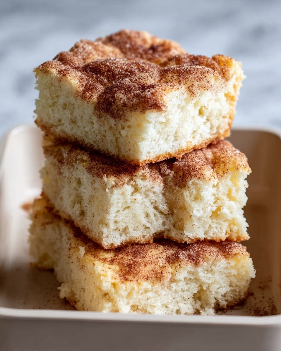The image shows a stack of three thick squares of soft, fluffy cake with a golden-brown crust on top and a light, airy inside. The top layer has a rough texture with a cinnamon sugar coating that looks slightly crispy, giving a slightly speckled look with darker brown spots. The cake squares are arranged in a white tray with a smooth edge, and the background has a white marbled texture. One square is placed on top of two others, showing the soft crumb inside clearly. photo taken with an iphone --ar 4:5 --v 7