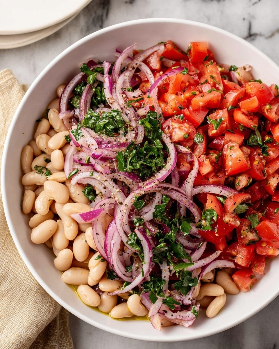 A white bowl filled with a fresh bean salad showing three main layers: the bottom layer is white cannellini beans with a smooth, slightly shiny texture; the middle layer consists of chopped bright red tomatoes cut into small chunks; the top layer has thin, curly slices of purple-red onion mixed with chopped green parsley leaves, all lightly sprinkled with black pepper and tiny oil droplets giving a glossy look. The bowl is placed on a white marbled surface with a beige cloth and a white plate partially visible in the background. photo taken with an iphone --ar 4:5 --v 7