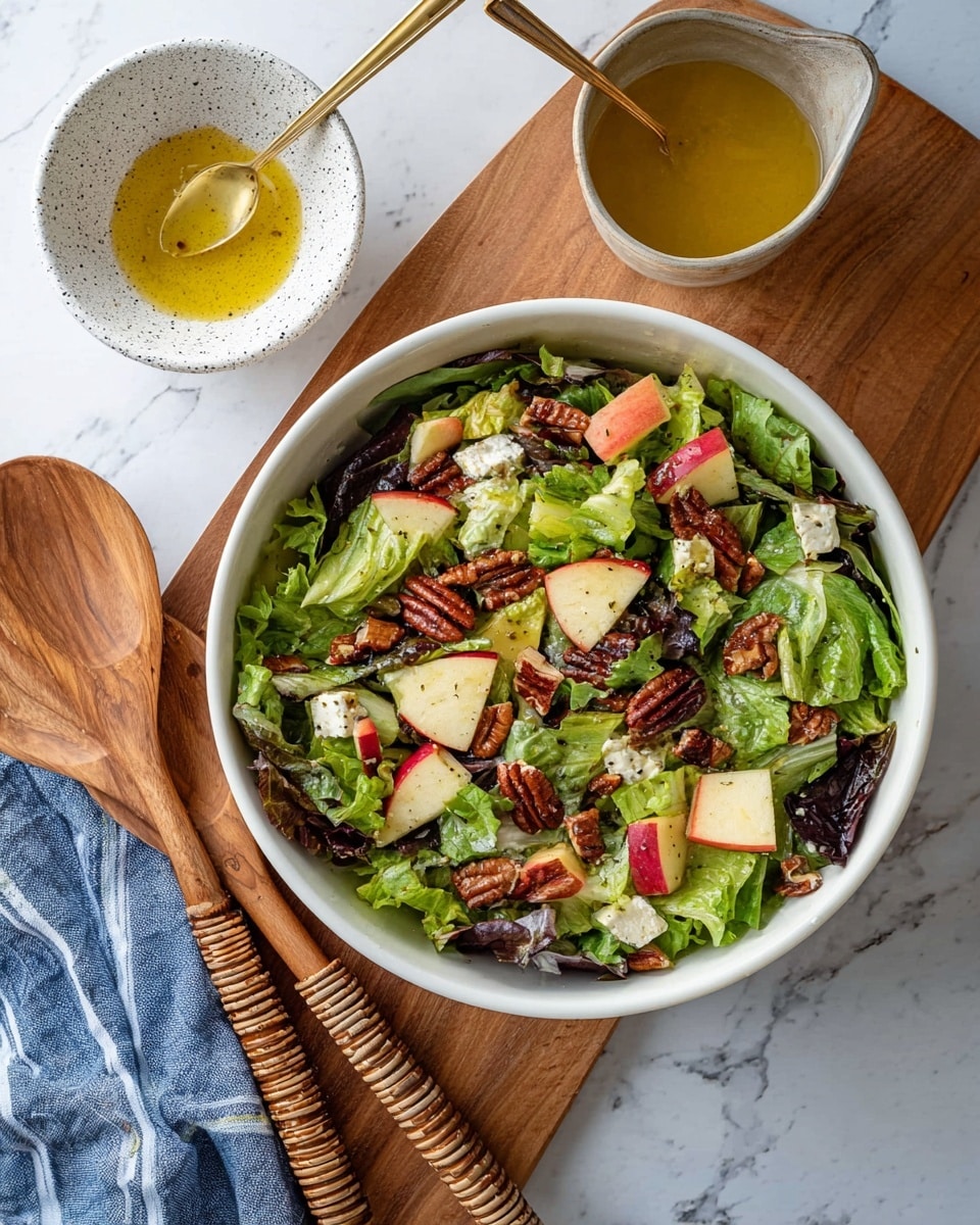 A white bowl filled with a fresh mixed salad sits on a wooden board over a white marbled surface. The salad has several layers: a base of green and dark leafy lettuce, scattered pieces of red apple with skin on, small chunks of white cheese, and crunchy brown pecans teasing the texture. Next to the bowl, a small white speckled bowl holds a yellow olive oil dressing with a golden spoon. Nearby, two wooden salad spoons with woven handles rest on a blue cloth with white stripes. photo taken with an iphone --ar 4:5 --v 7