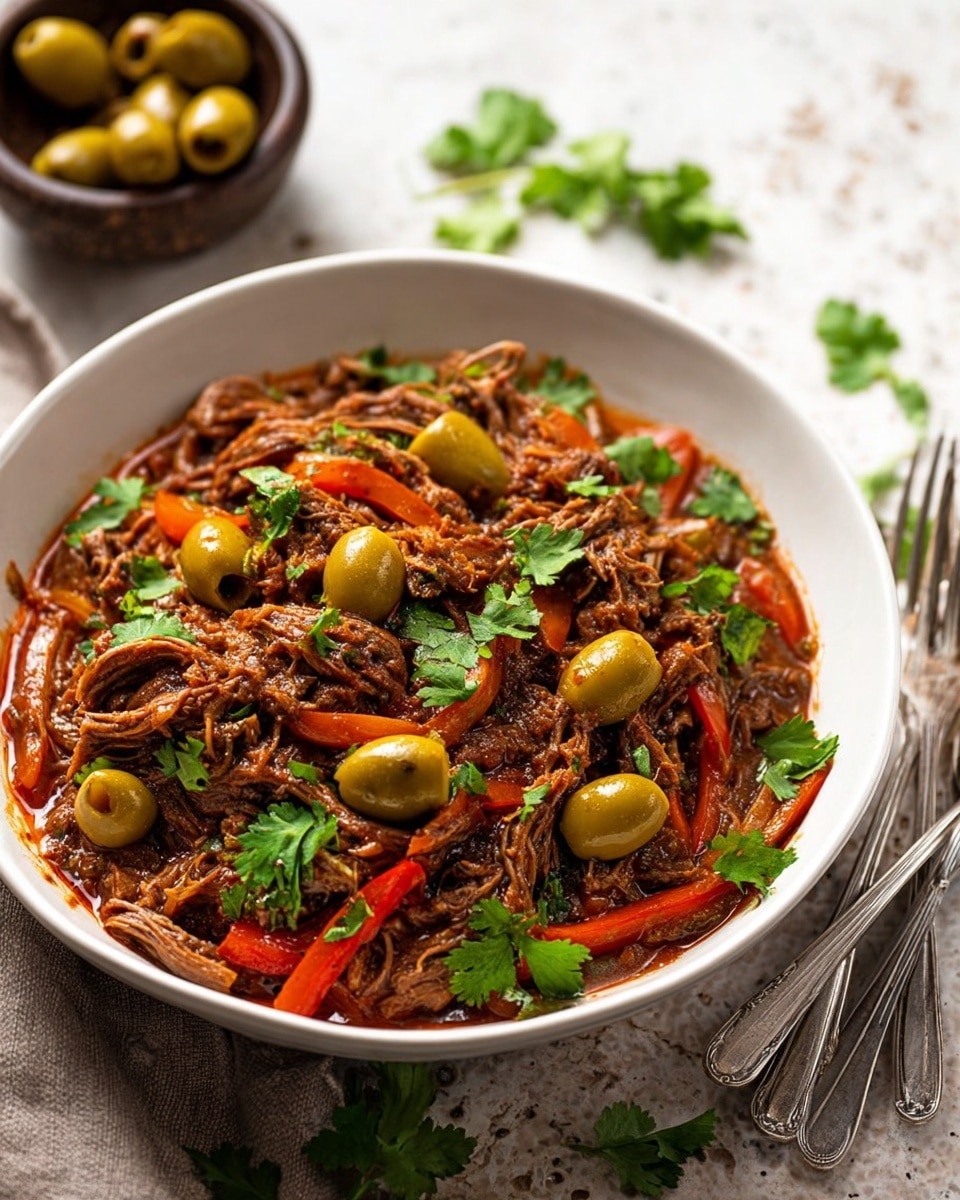 A white bowl filled with shredded brown meat mixed evenly with green olives and thin strips of red and orange peppers, all coated lightly in a rich reddish-brown sauce. The dish is topped with fresh green cilantro leaves scattered over the surface. The bowl rests on a white marbled surface, with a few sprigs of cilantro nearby and a stack of forks and spoons blurred in the background. photo taken with an iphone --ar 4:5 --v 7