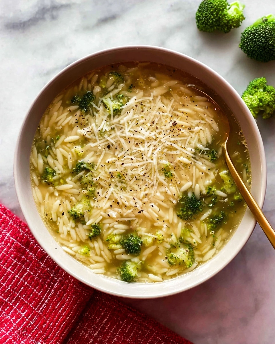 A white bowl filled with clear broth soup with small orzo pasta pieces scattered throughout, mixed with small green broccoli florets. The top layer is sprinkled with shredded white cheese and a dusting of black pepper. A shiny gold spoon is partially submerged in the soup, resting on the right side of the bowl. The bowl sits on a white marbled surface with three small broccoli florets placed near the top right corner and a red and white striped cloth partially visible at the bottom left. photo taken with an iphone --ar 4:5 --v 7