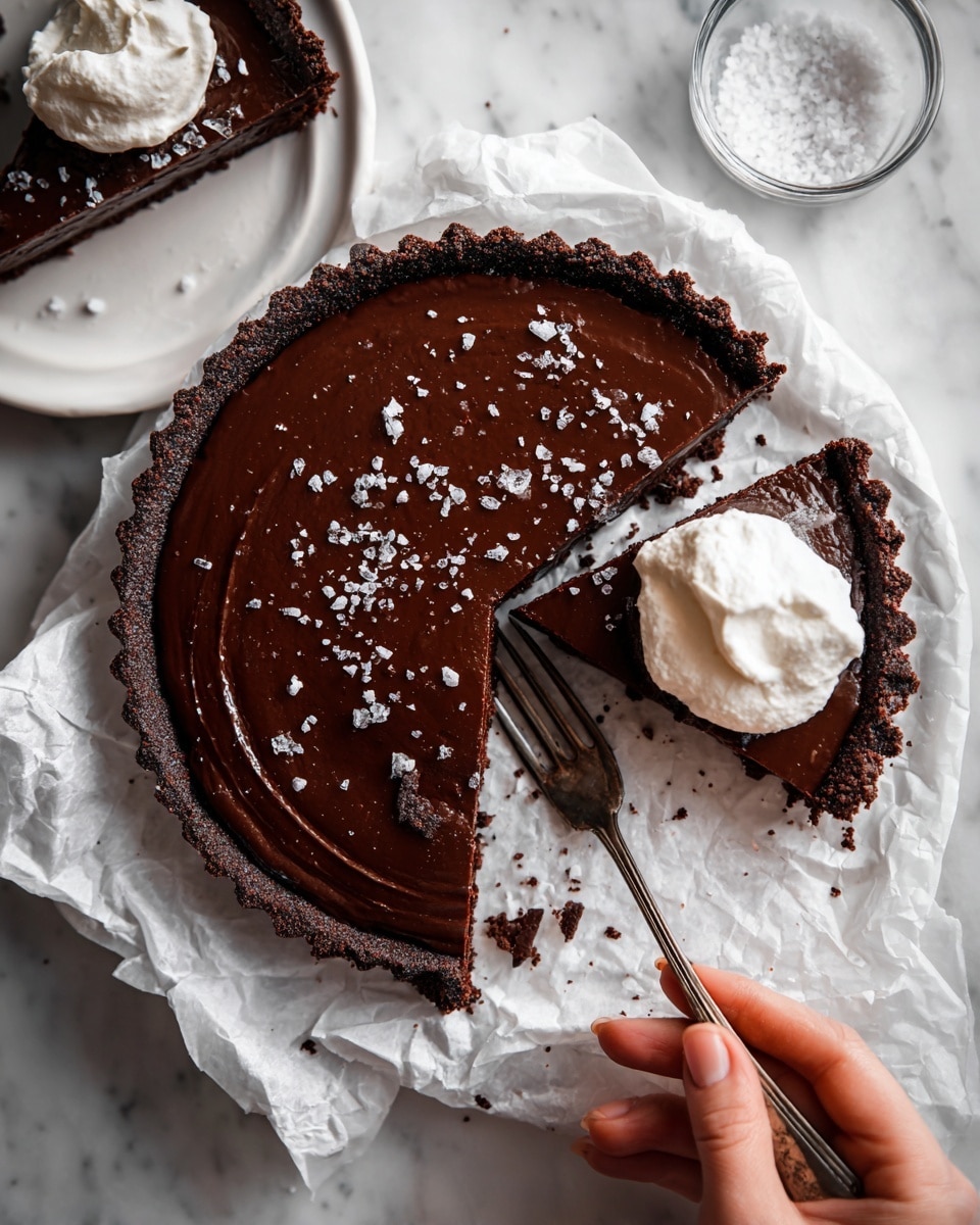 A dark chocolate tart with two visible layers sits on crumpled white paper over a white marbled surface. The bottom layer is a dark, crumbly chocolate crust with a rough texture around the edges. The top layer is a smooth, glossy dark chocolate filling, sprinkled with coarse white sea salt. One slice is cut out from the tart, and a slice is placed on a white plate to the upper left, partially eaten with a silver fork. Another slice is on crumpled white paper at the bottom, topped with a fluffy white cream. A woman's hand holds a fork near the cream-covered slice. A small clear glass bowl with white salt is to the right of the tart. Photo taken with an iphone --ar 4:5 --v 7
