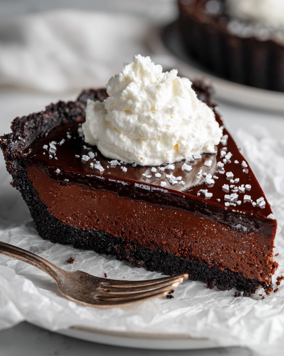 The image shows a slice of chocolate tart on crumpled white paper over a white marbled surface, with a silver fork holding a piece of the tart topped with a dollop of white whipped cream. The tart has a dark, crumbly crust base, a thick middle layer of dense chocolate filling with a smooth and creamy texture, and a glossy, dark chocolate ganache layer on top sprinkled with small white flakes. The whipped cream on the fork is fluffy and soft, sitting on the rich chocolate piece. Photo taken with an iphone --ar 4:5 --v 7
