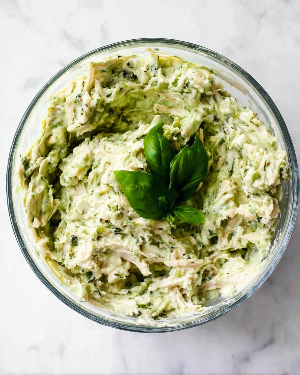 A clear glass bowl holds a creamy mixture with a thick texture, mainly light green with visible small white and darker green bits. The dish has shredded pieces scattered throughout, giving a fibrous look. There is a small cluster of fresh bright green basil leaves placed on top, adding a fresh touch. The bowl is on a white marbled surface. photo taken with an iphone --ar 4:5 --v 7
