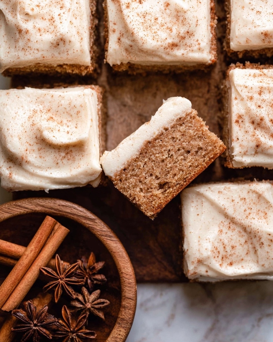 The image shows a close-up view of square pieces of spiced cake arranged closely together, each topped with a thick layer of smooth, creamy white frosting that looks soft and slightly swirled. The frosting has a light dusting of cinnamon powder on top, adding a warm brown speckled texture. One piece is held slightly above the other cake squares, showing the inside texture of the cake, which is light brown, moist, and fluffy with visible crumb structure. Next to the cake, there is a wooden bowl filled with whole cinnamon sticks and star anise, set on a surface with a white marbled texture. photo taken with an iphone --ar 4:5 --v 7