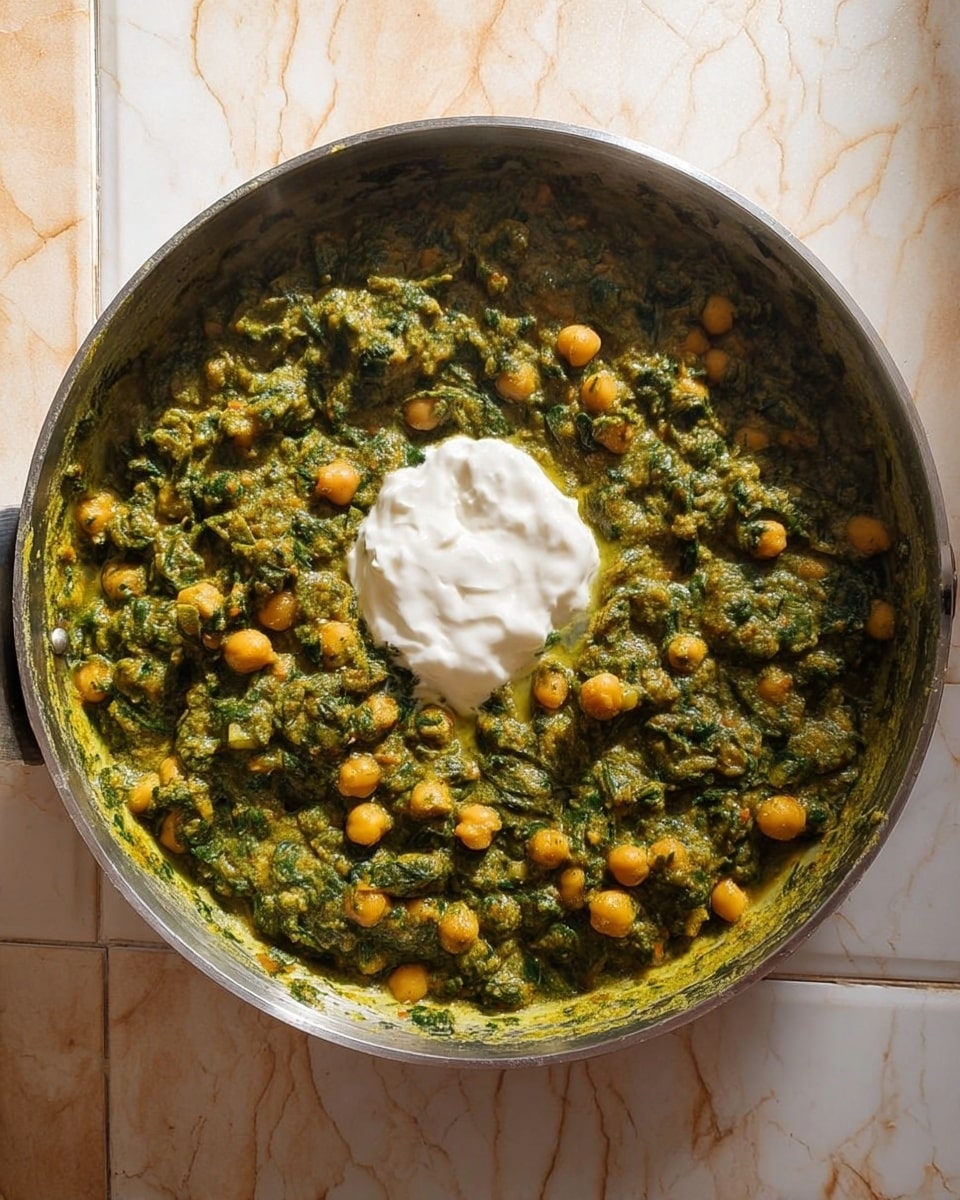 A metal pan filled with a thick green spinach-based curry mixed with yellow chickpeas, showing a dense, textured sauce with small bits of spinach and chickpeas spread evenly throughout. In the center, there is a dollop of smooth, creamy white yogurt placed on top of the curry, adding contrast to the green and yellow hues. The pan sits on a white marbled surface with light patterns. The background shows parts of tiled walls in soft beige tones. photo taken with an iphone --ar 4:5 --v 7