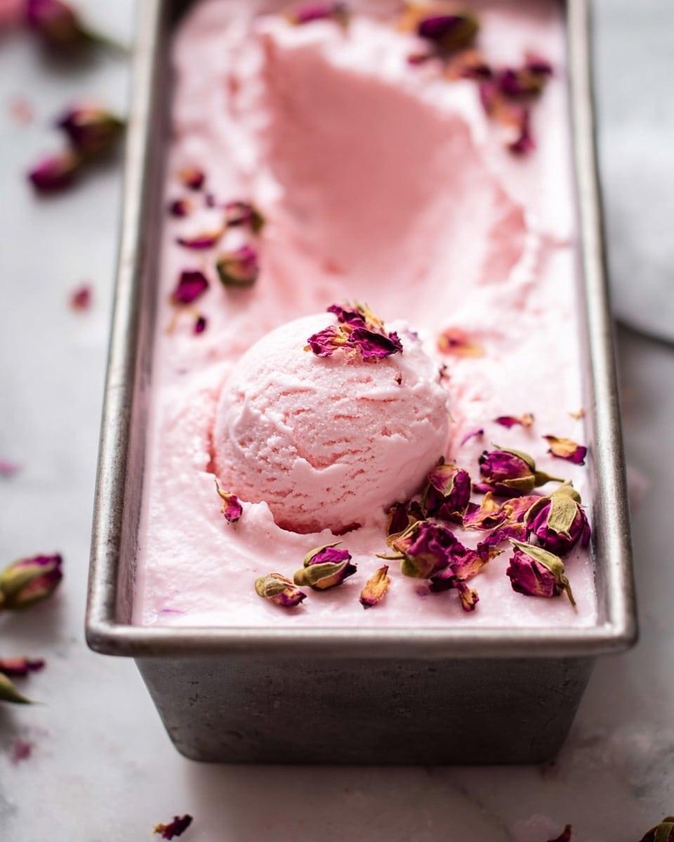 A metal rectangular container holds one thick layer of light pink ice cream with a smooth, whipped texture. On top, small dried rose petals and buds in deep purple and dusty pink are scattered evenly, adding color contrast. In the center, a scoop of the same pink ice cream is neatly placed, topped with more dried rose petals. The scene is set on a white marbled surface. photo taken with an iphone --ar 4:5 --v 7
