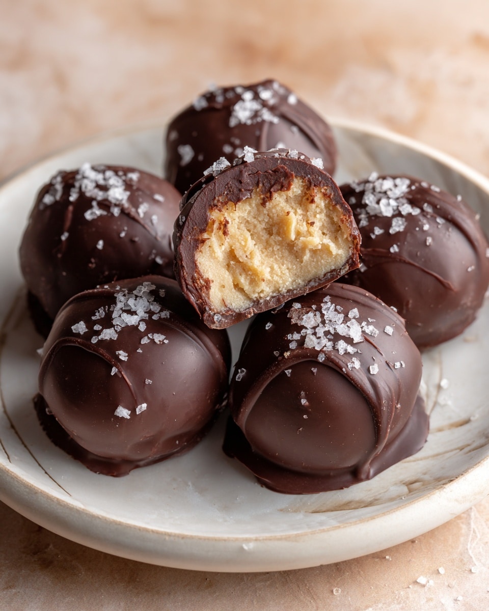The image shows five small round treats covered in dark chocolate with a smooth and shiny surface, arranged on a white plate with a white marbled texture underneath. One treat is cut open, showing a thick, creamy light tan filling inside with a soft texture. Some granules of coarse salt are sprinkled on top of the chocolate coating. The lighting highlights the glossy chocolate and soft filling clearly, with a warm tone in the background. photo taken with an iphone --ar 4:5 --v 7