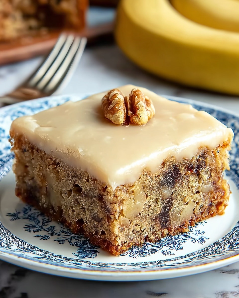 A square slice of cake with two visible layers sits on a white plate with blue patterns. The bottom layer is light brown with dark spots, showing a crumbly texture with bits of fruit or nuts inside. The top layer is a smooth, light beige frosting with a glossy finish, topped with a single walnut piece in the center. The sides reveal a moist, dense cake with a slightly crunchy base. The plate rests on a white marbled surface with a blurred banana and another fork in the background. photo taken with an iphone --ar 4:5 --v 7