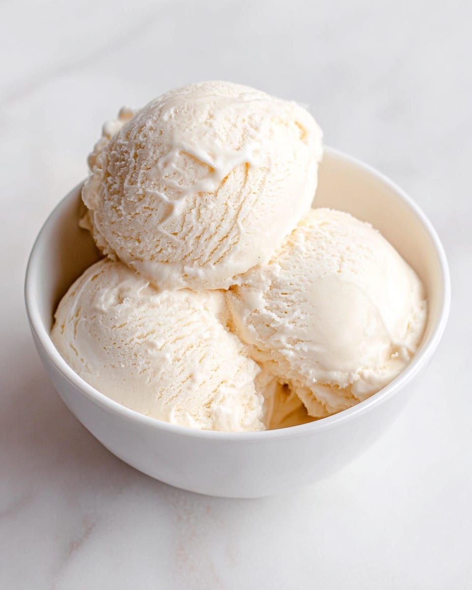 A white bowl filled with three scoops of creamy vanilla ice cream. Each scoop has a smooth but slightly textured surface with small ridges and soft folds, showing the rich, dense texture of the ice cream. The bowl sits on a white marbled surface, and the ice cream appears slightly glossy with a soft, off-white color. photo taken with an iphone --ar 4:5 --v 7