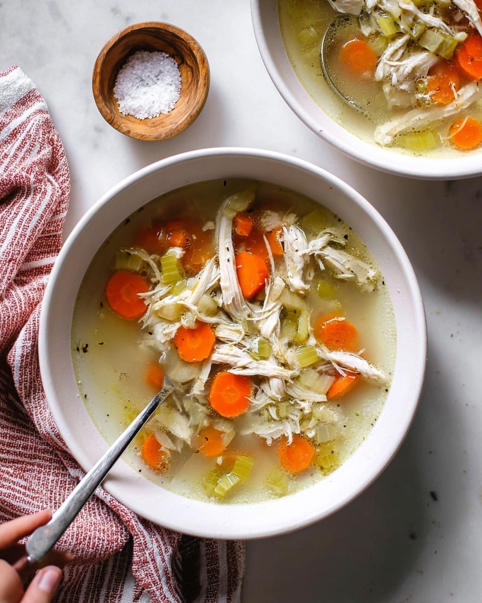 A white bowl filled with clear chicken soup that has shredded white chicken pieces spread evenly throughout. Bright orange carrot slices and pale green celery chunks are mixed in, floating in the light broth. A silver spoon rests inside the bowl, partially submerged in the soup. The bowl sits on a white marbled surface next to a small wooden bowl with coarse salt, and a woman's hand is holding a red and white striped cloth nearby. Another bowl of the same soup is partially visible in the top right corner. Photo taken with an iphone --ar 4:5 --v 7