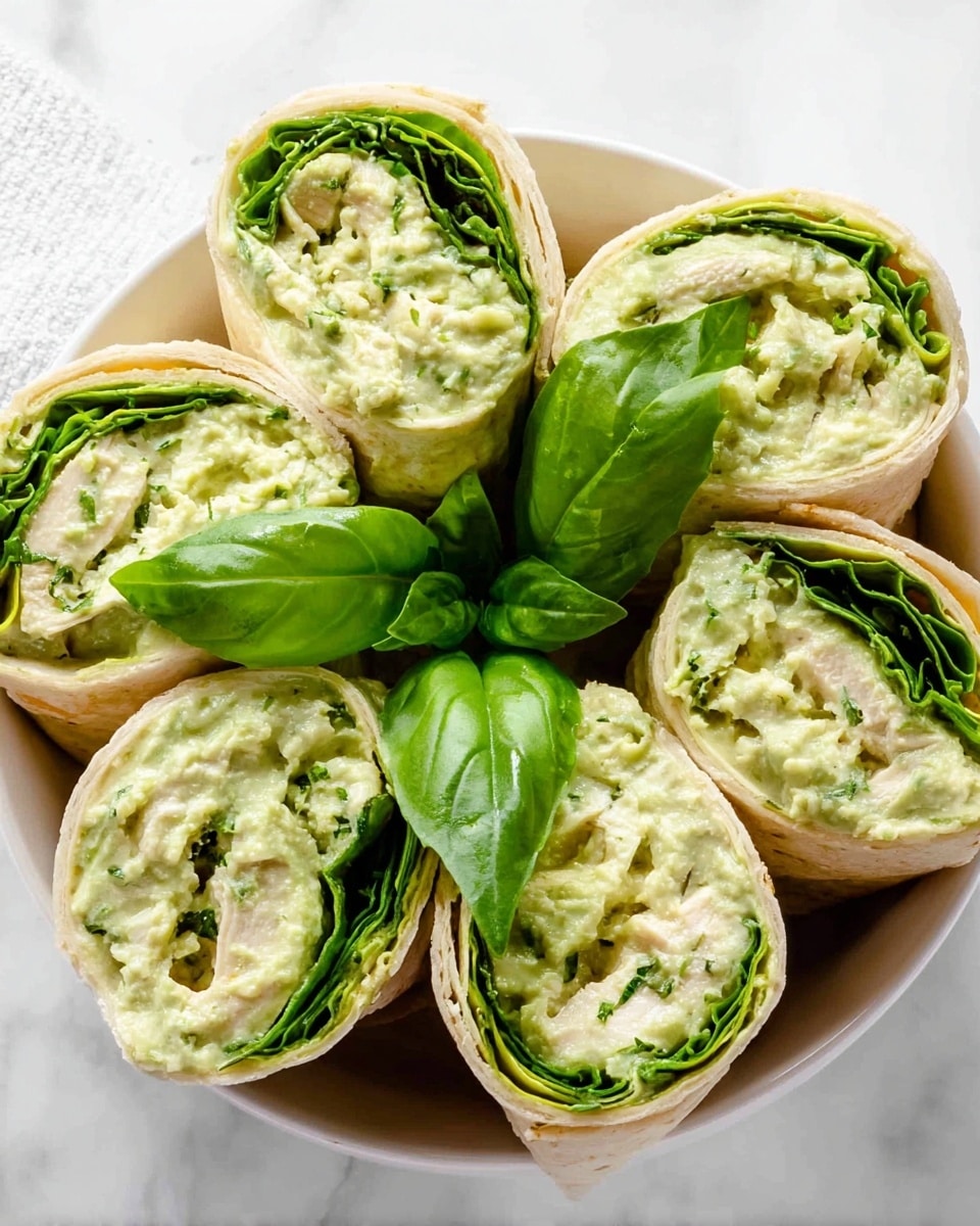The image shows six tortilla roll-ups arranged in a white bowl on a white marbled surface. Each roll-up has three visible layers: the outer pale beige tortilla wrap, a middle layer of bright green fresh spinach leaves, and an inner creamy, light green filling that looks smooth with small chunks, likely a pesto chicken salad mixture. The roll-ups are cut at an angle to show the layered filling inside. In the center of the bowl, there are several fresh basil leaves adding a vibrant green contrast. The overall look is fresh, creamy, and green with textures from soft tortillas, leafy spinach, and creamy salad. Photo taken with an iphone --ar 4:5 --v 7