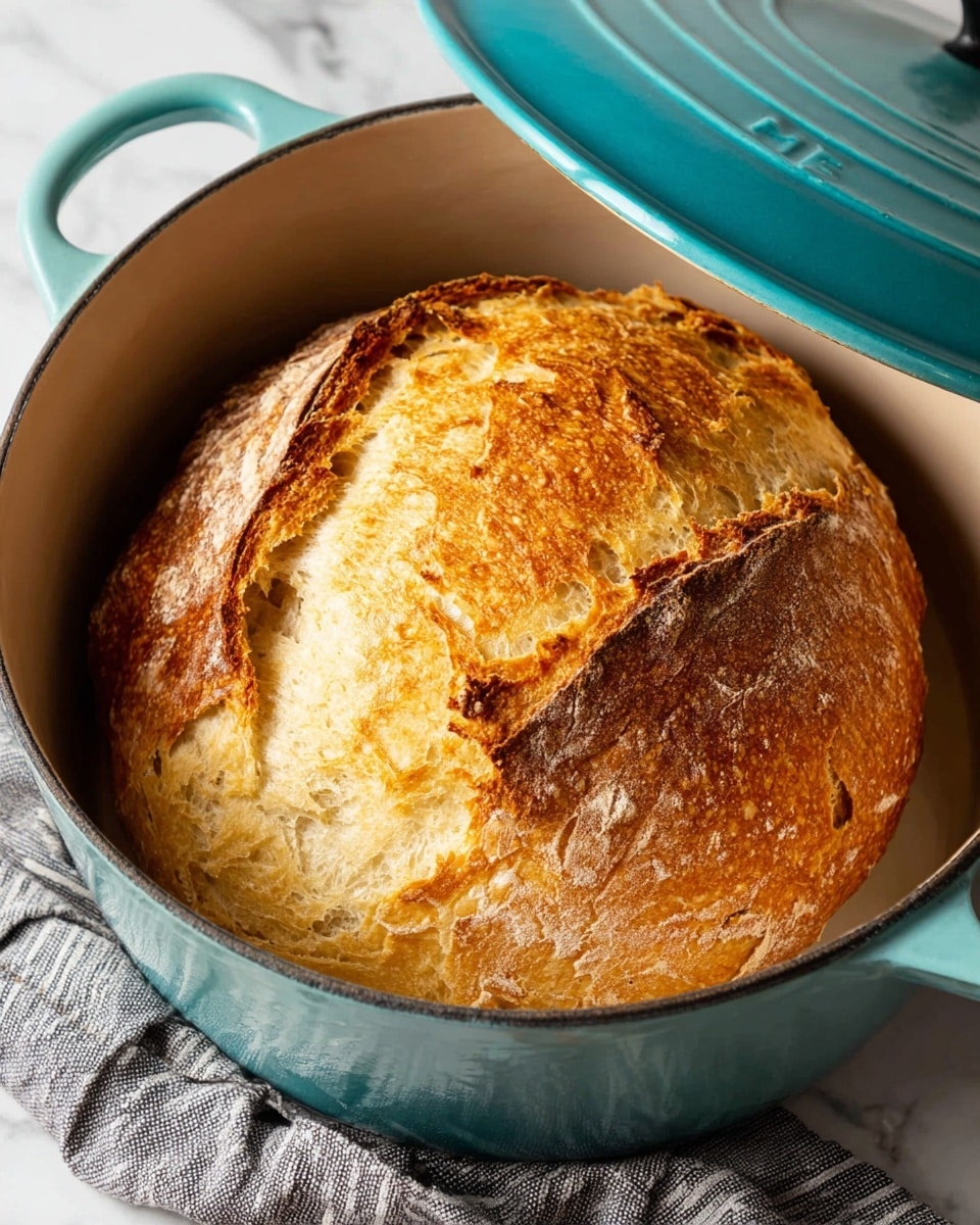 A round loaf of bread inside a white enamel pot with a turquoise lid that is tilted open, showing the bread's golden brown crust with deep cracks and a rough, textured surface. The bread dome rises above the pot's rim, with a crisp and slightly uneven crust in varying shades of golden yellow and brown. The pot rests on a white marbled surface with a gray striped cloth beside it. Photo taken with an iphone --ar 4:5 --v 7