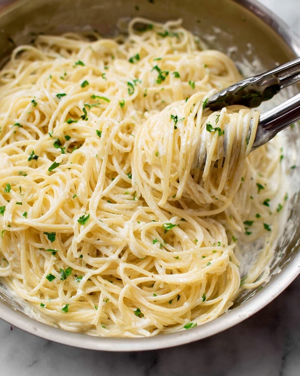 A close-up view of cooked spaghetti pasta in a round metal pan, coated evenly with a creamy white sauce. The noodles form a soft, tangled mass with a smooth and slightly shiny texture, lightly sprinkled with small bits of fresh green herbs scattered on top. A pair of silver tongs are lifting some pasta from the pan, showing the long, thin strands stretching slightly. The background shows a white marbled texture. photo taken with an iphone --ar 4:5 --v 7