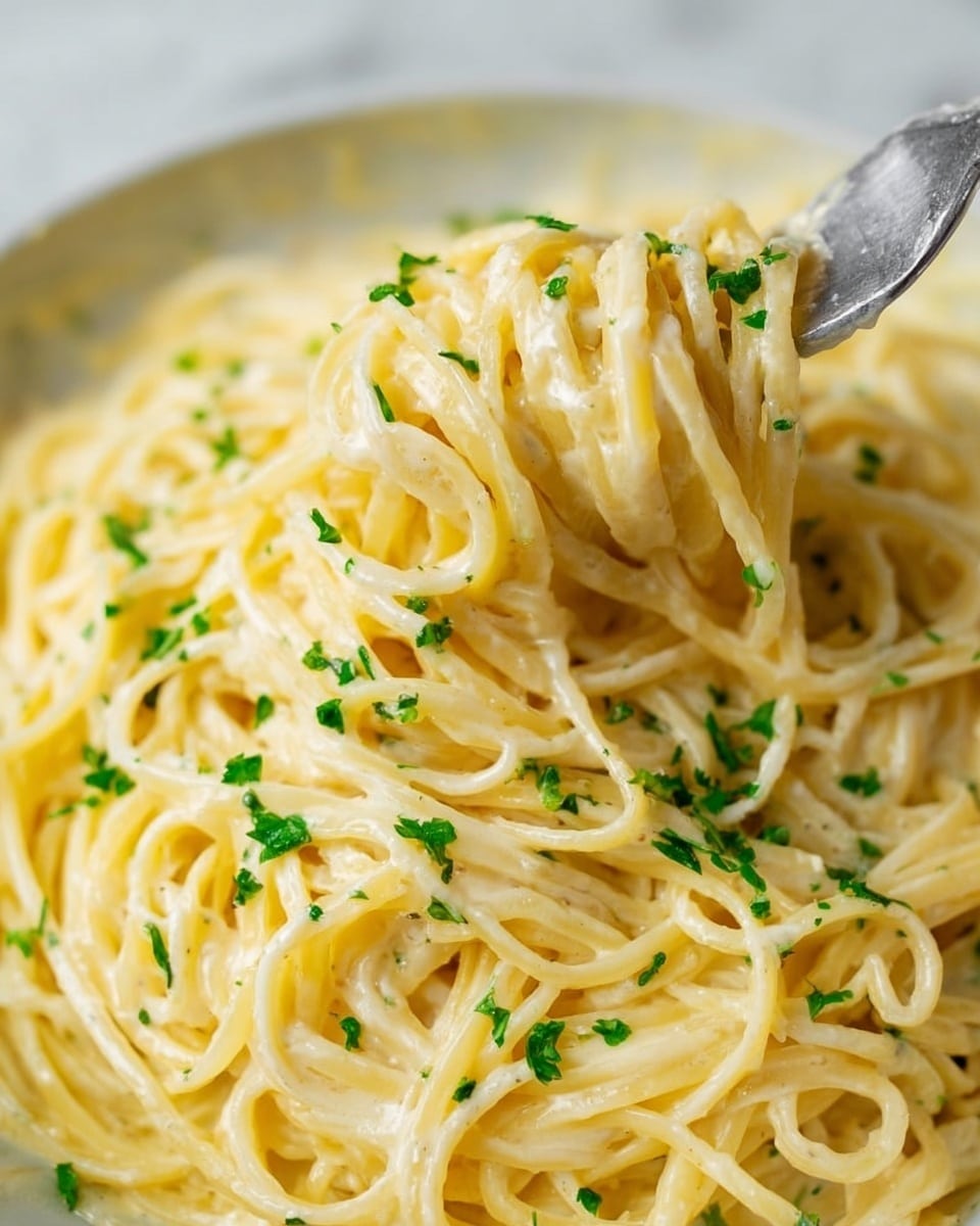 A close-up view of creamy spaghetti pasta tossed in a smooth, pale yellow sauce with a glossy texture, sprinkled with small, bright green chopped herbs scattered evenly on top. The pasta strands are thick and well coated, twisted and heaped naturally, with a silver fork lifting a portion from the right side of the image. The dish is set against a white marbled background. photo taken with an iphone --ar 4:5 --v 7